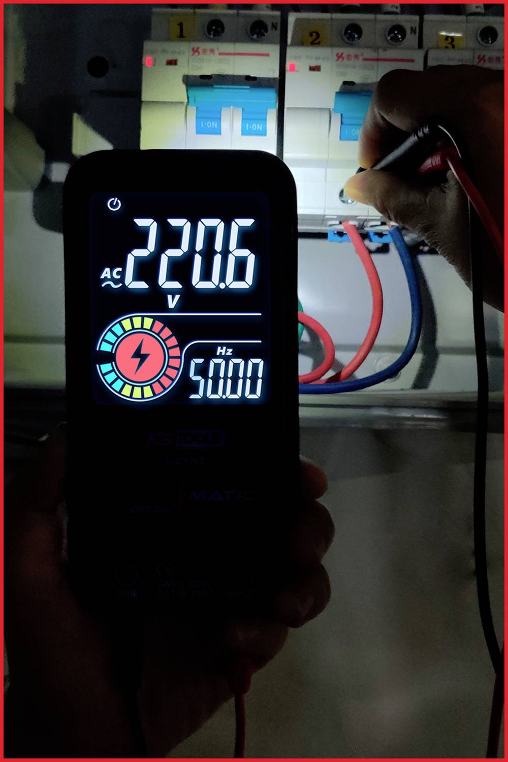 En person måler 220,6 V AC ved 50,0 Hz med et digitalt voltmeter ved en åben sikringsboks med tilsluttede ledninger.