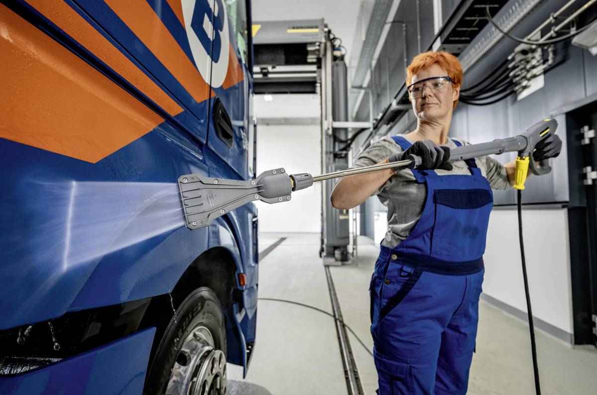 En person iført beskyttelsesudstyr power washer en stor køretøj i et industrielt miljø. Køretøjet er blåt med orange detaljer.