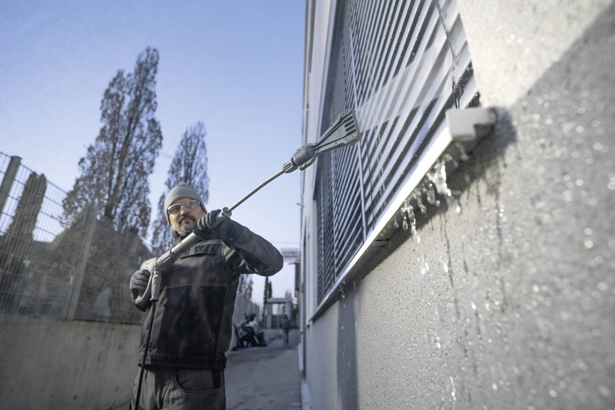 En person i varmt tøj rengør udvendige vinduesjalusier på en bygning med en langskaftet børste, vand der sprøjter, på en solrig dag.