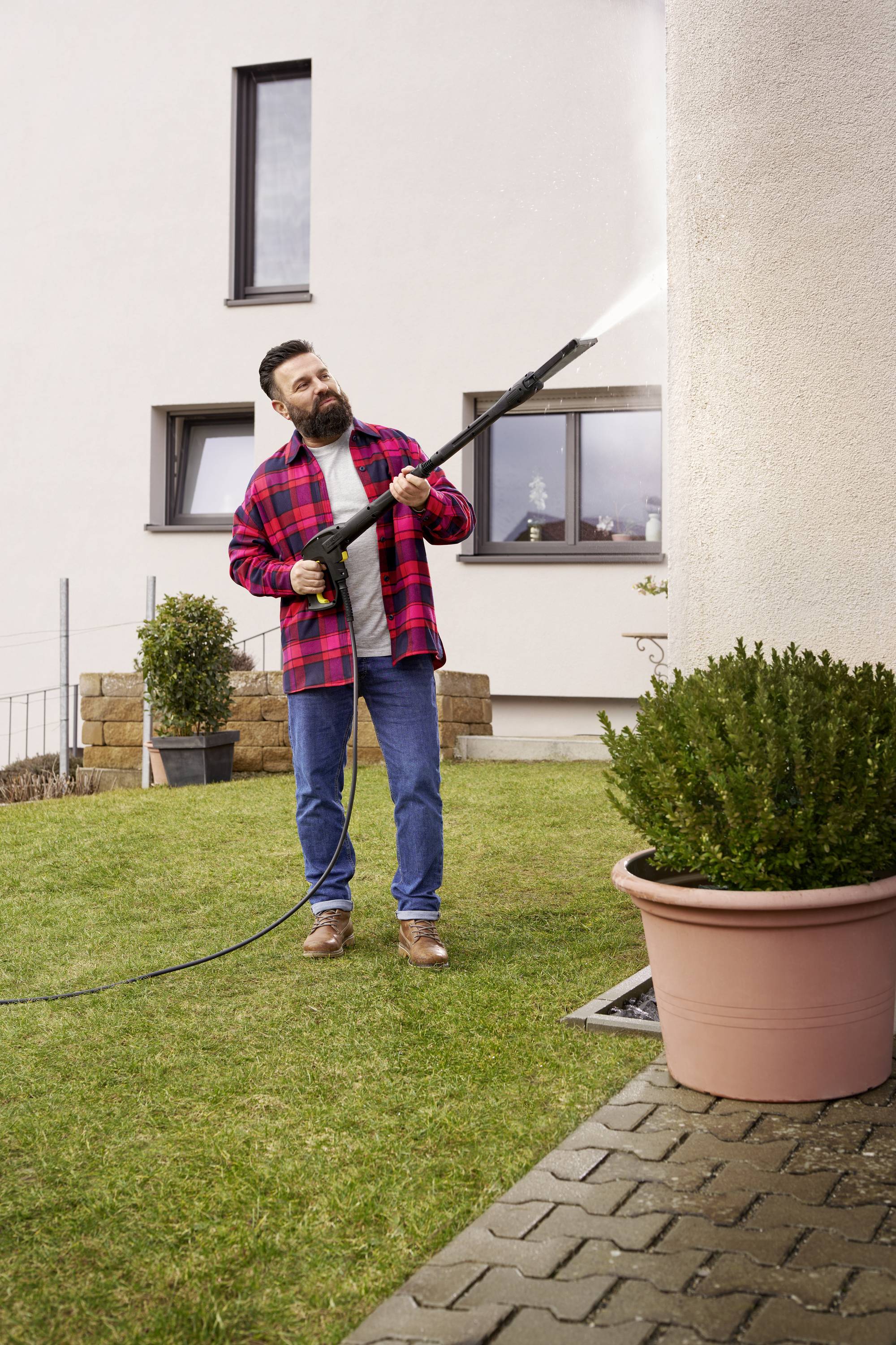 En mand rengør husets facade med en højtryksrenser, omgivet af en have med plantekasser og græsplæne.