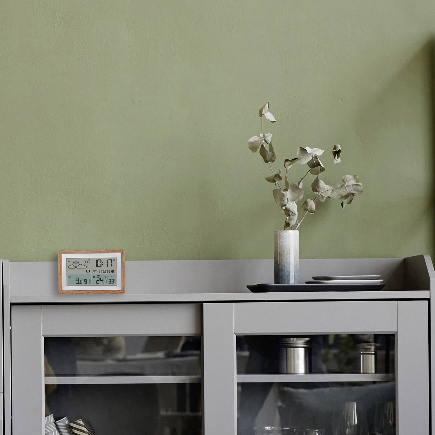 Et gråt møbel foran en grøn væg, hvorpå der står en vase med tørrede blomster og en digital kalender.