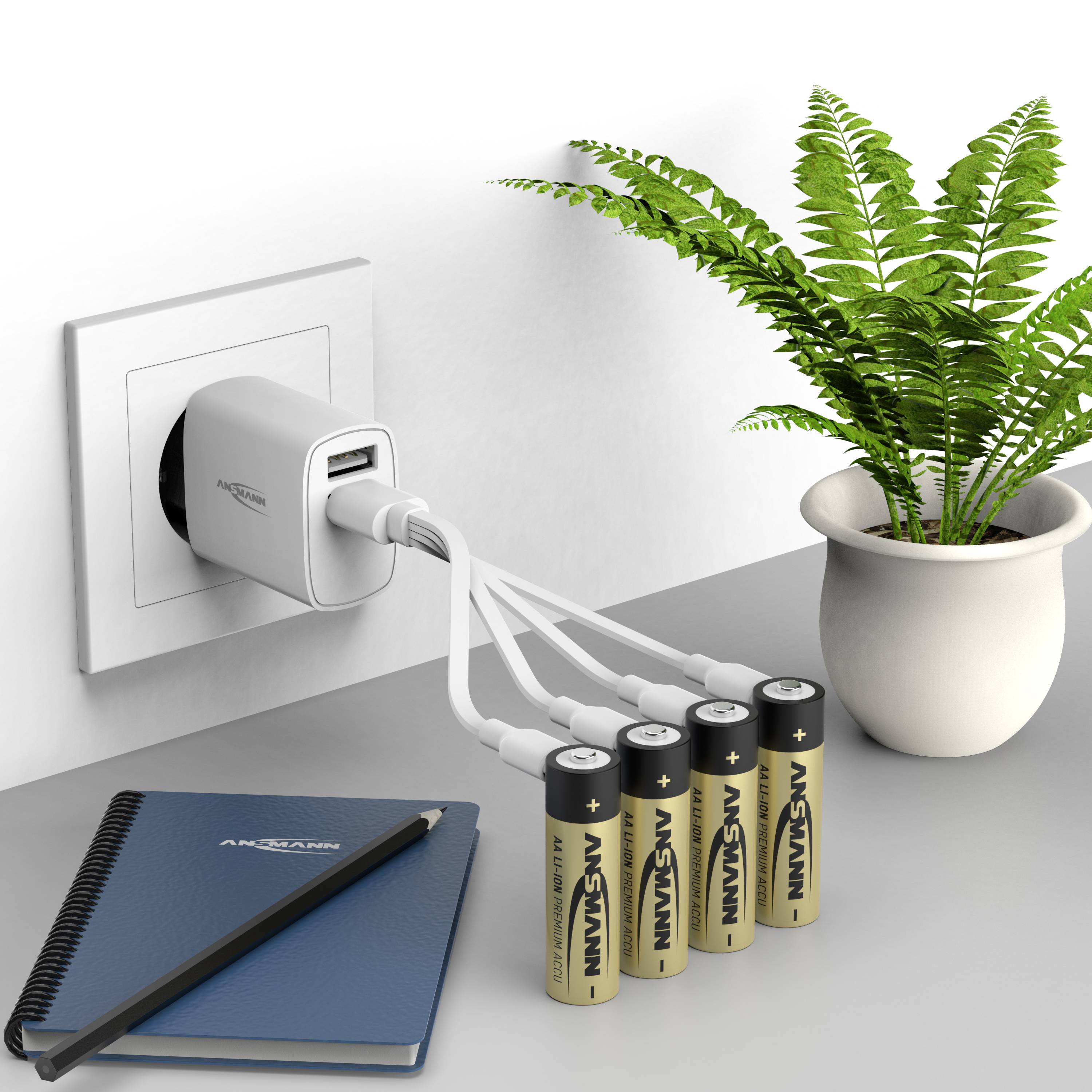 Et stik med en USB-oplader oploader fire AA-batterier. Ved siden af står en grøn plante i en potte og en notesbog på bordet.