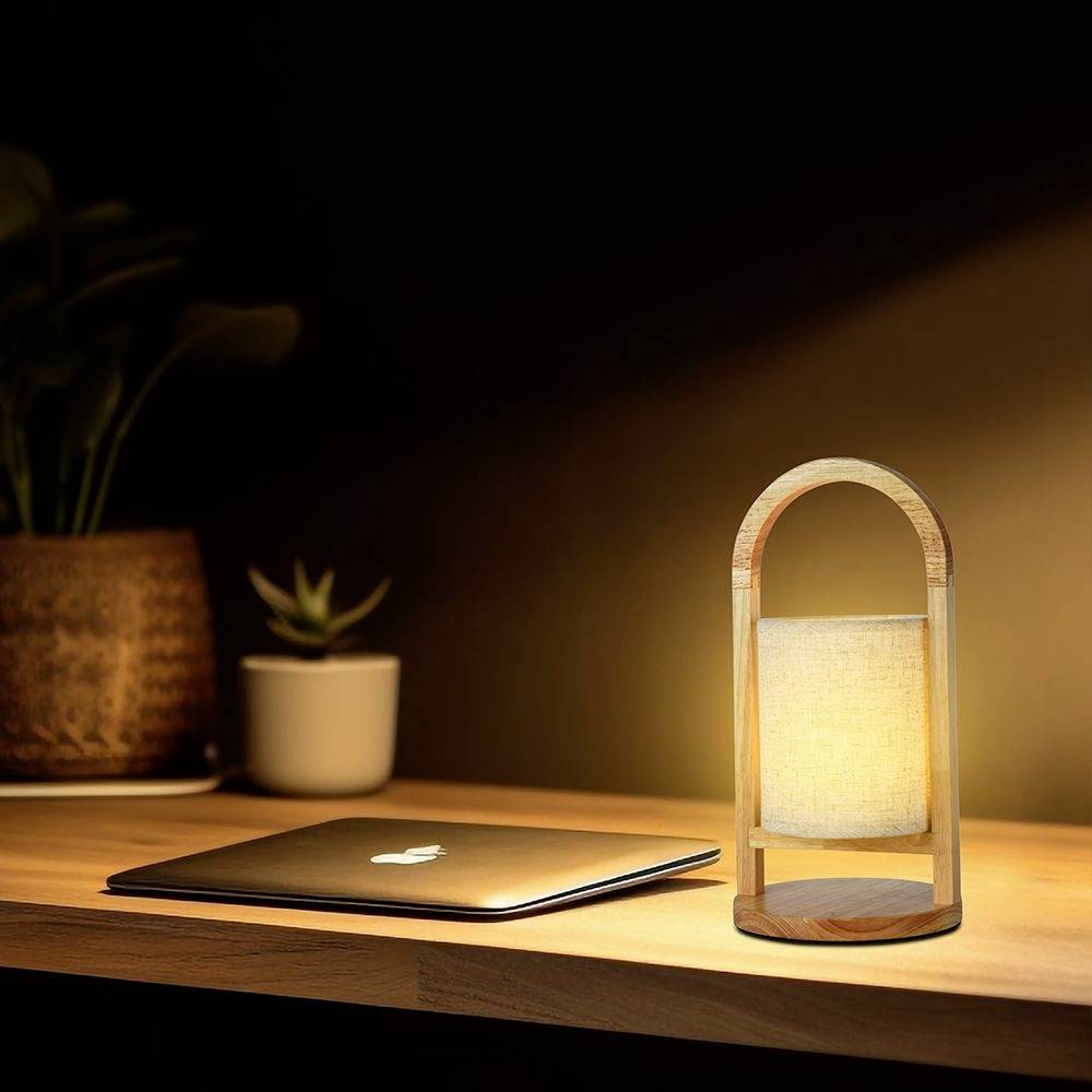 En bærbar computer ligger på et træbord ved siden af en lysende bordlampe, med en plante i baggrunden.