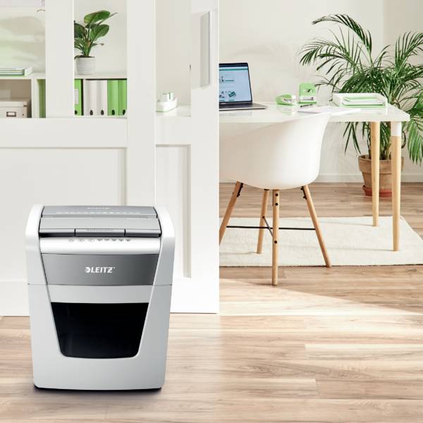 En makuleringsmaskine står i et moderne kontor. I baggrunden ses et skrivebord med stol, planter og en bærbar computer.