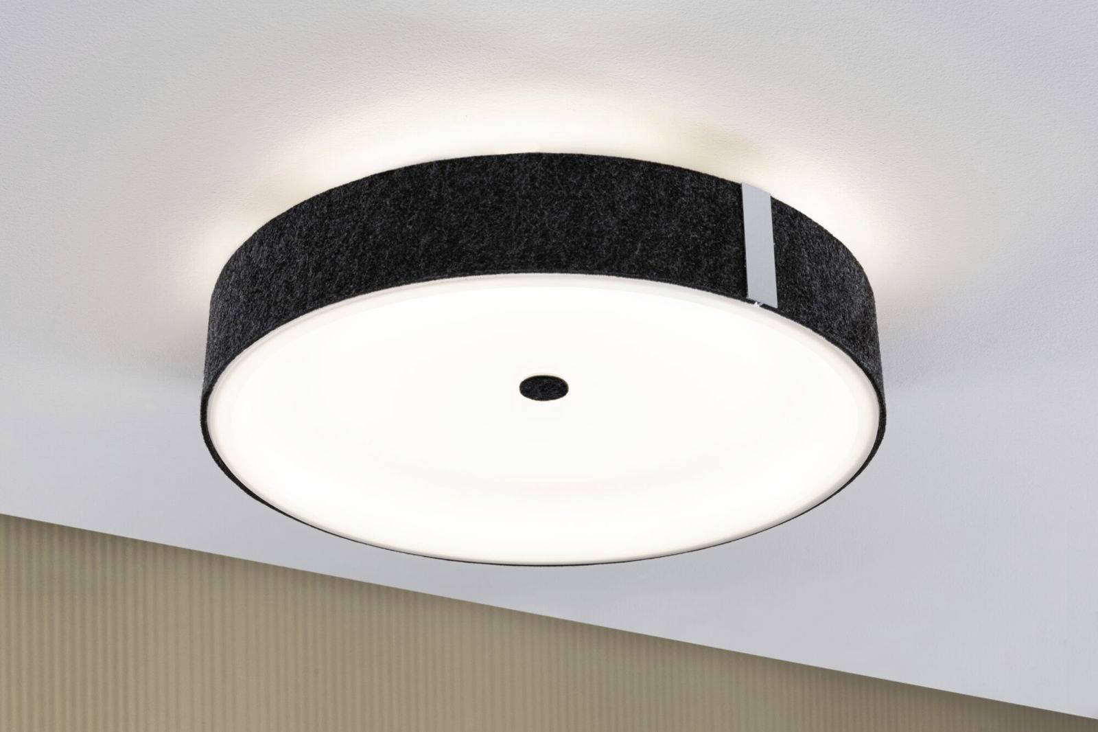 En rund, moderne loftslampe med sort kant lyser på en hvid lofts flade. Den udsender et diffust, ensartet lys.