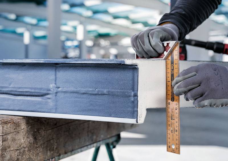 En person måler kanten af et stålbyggeelement ved hjælp af en vinkelmåler og et målebånd i et industrielt miljø.