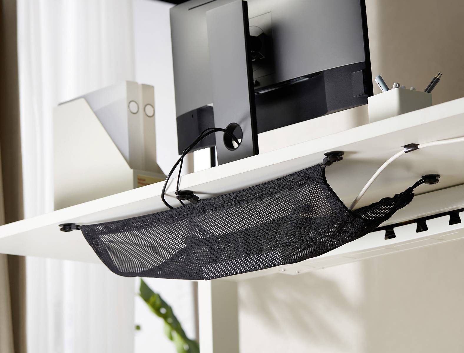 Et sort net under et skrivebord organiserer kabler. Oven på står en computer, ved siden af ligger dokumentmapper og pennen.