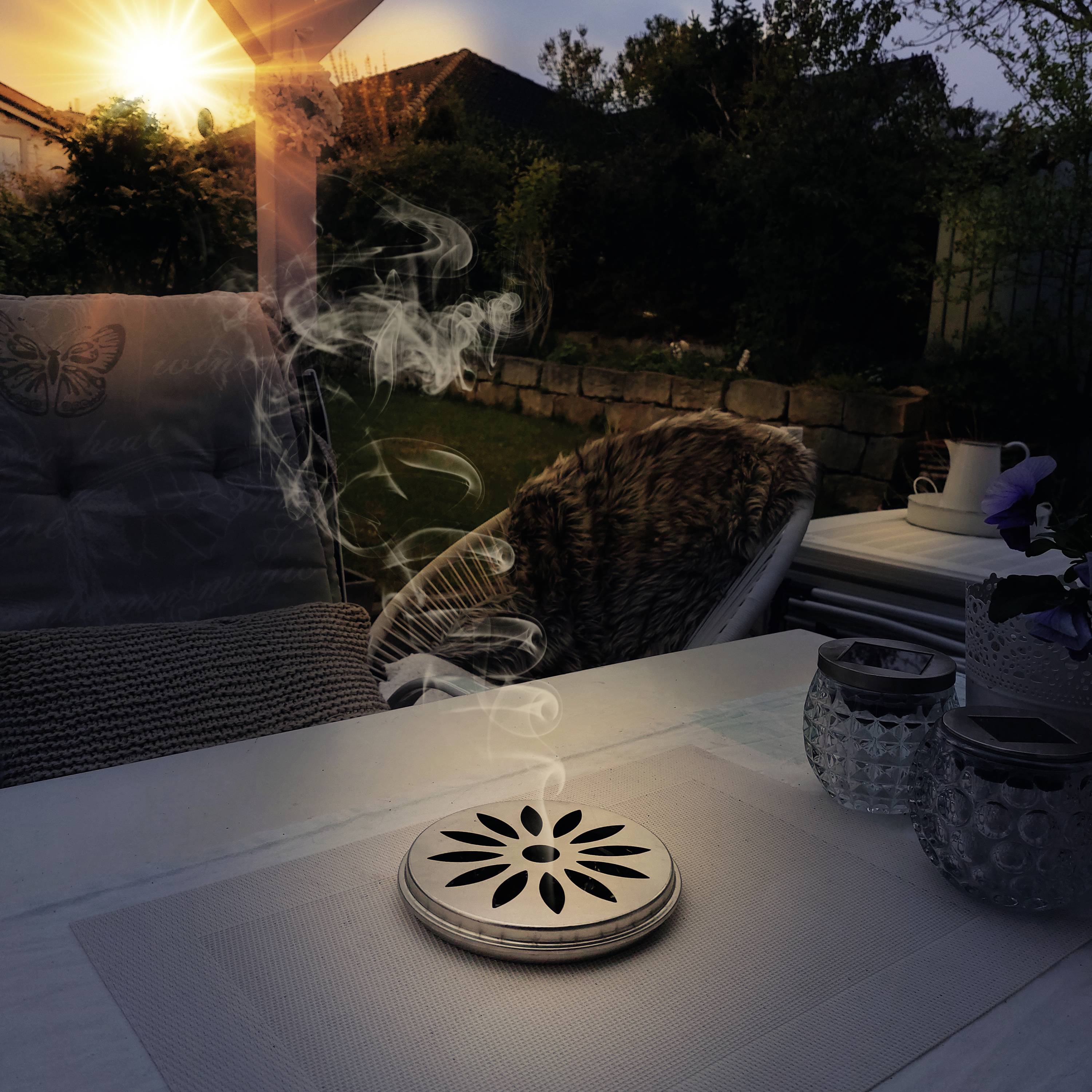 En hyggelig have ved solnedgang med en terrasse, hvor der står en dekorativ røgdiffuser, omgivet af planter.