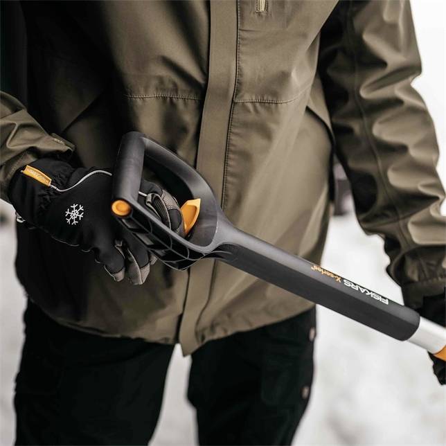 En person holder en Fiskars-skovl med sort greb i hånden. Personen bærer en jakke og handsker med snefnug-symboler.