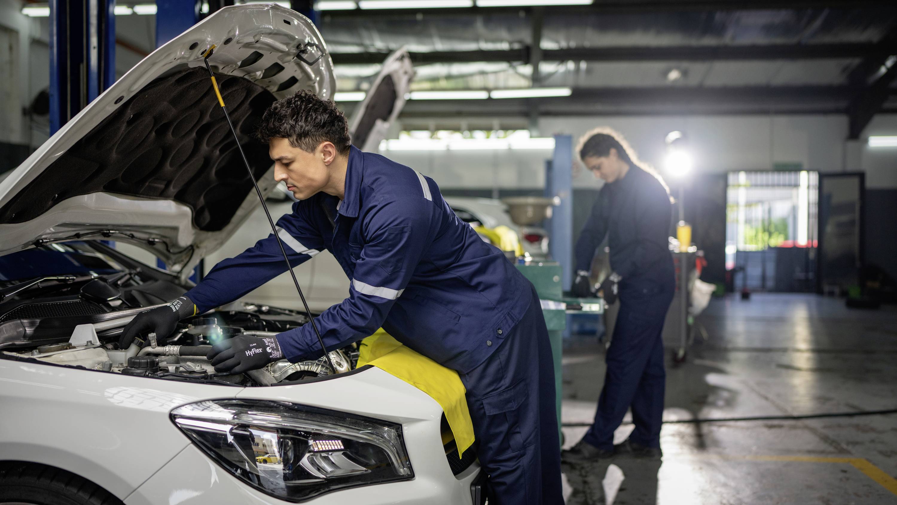 En mekaniker arbejder under motorhjelmen på en bil i et værksted, mens en anden mekaniker er beskæftiget i baggrunden.