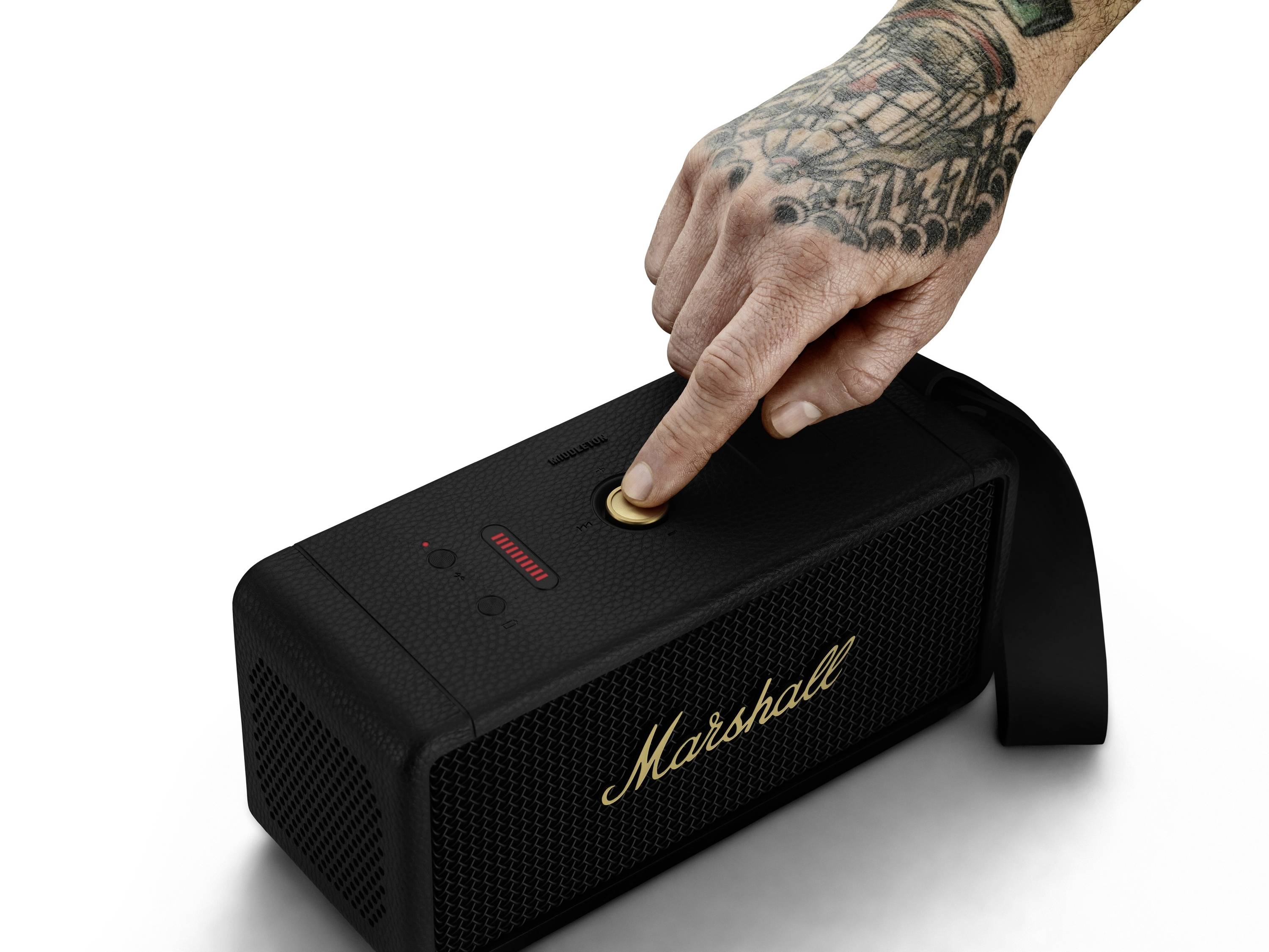 En tatoveret hånd trykker på tænd-knappen på en bærbar højtaler fra mærket Marshall. Højttaleren har et sort kabinet.