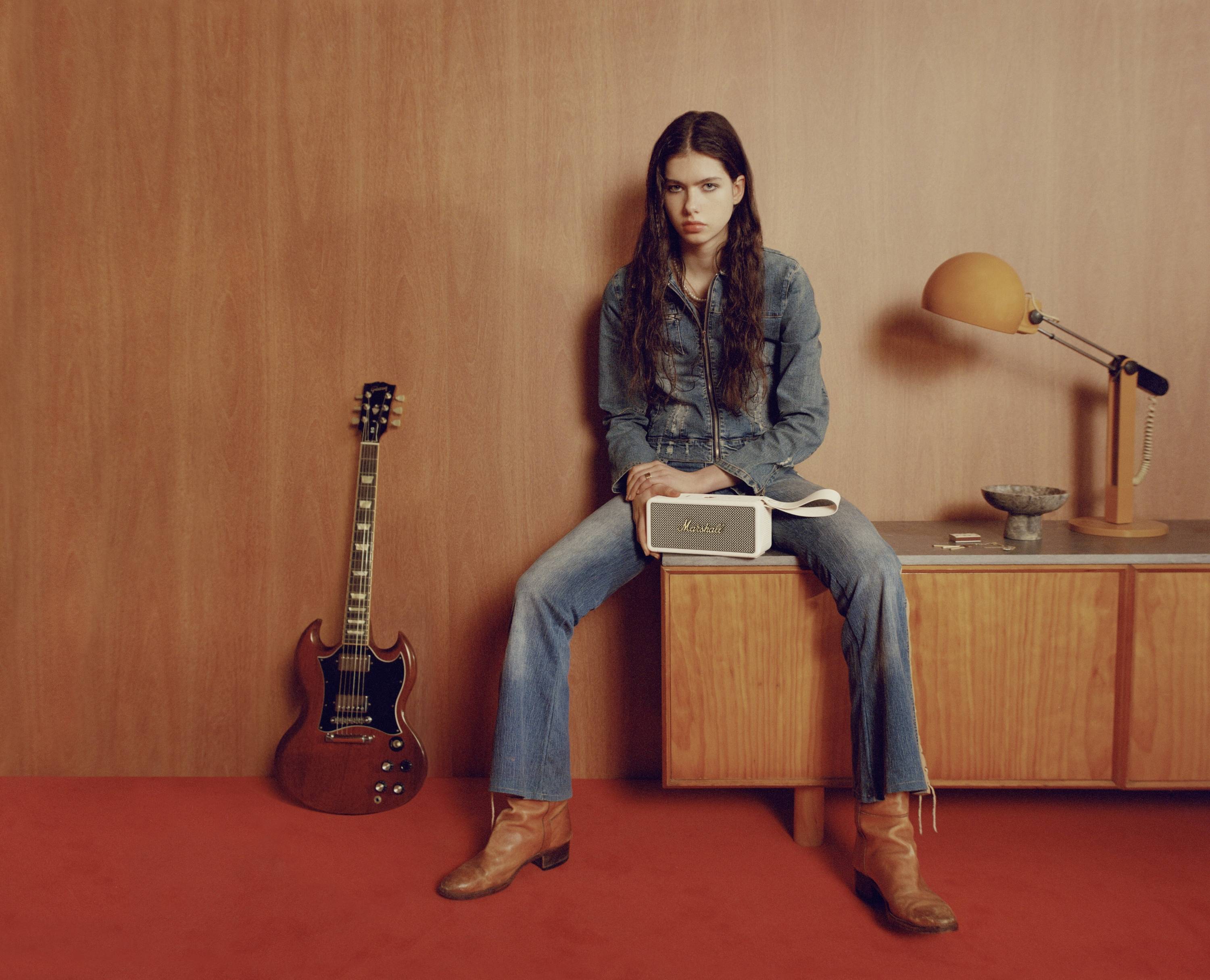 En person i jeans sidder på et sideboard ved siden af en guitar og en lampe, med en bog i hånden. Retro-stil.