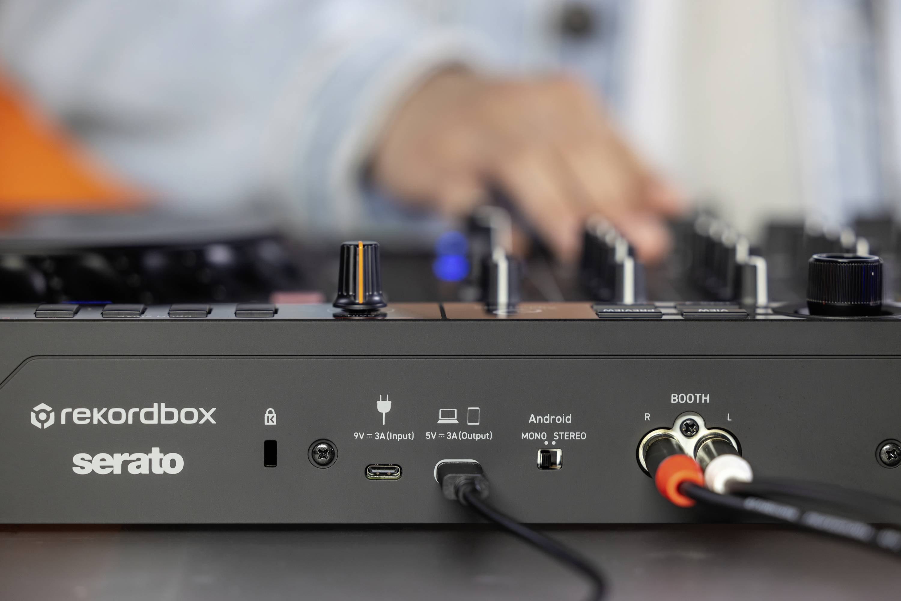 Nærbillede af et DJ-mixerpult med indgangs- og udgangsstik. En uskarp DJ betjener knapperne i baggrunden.