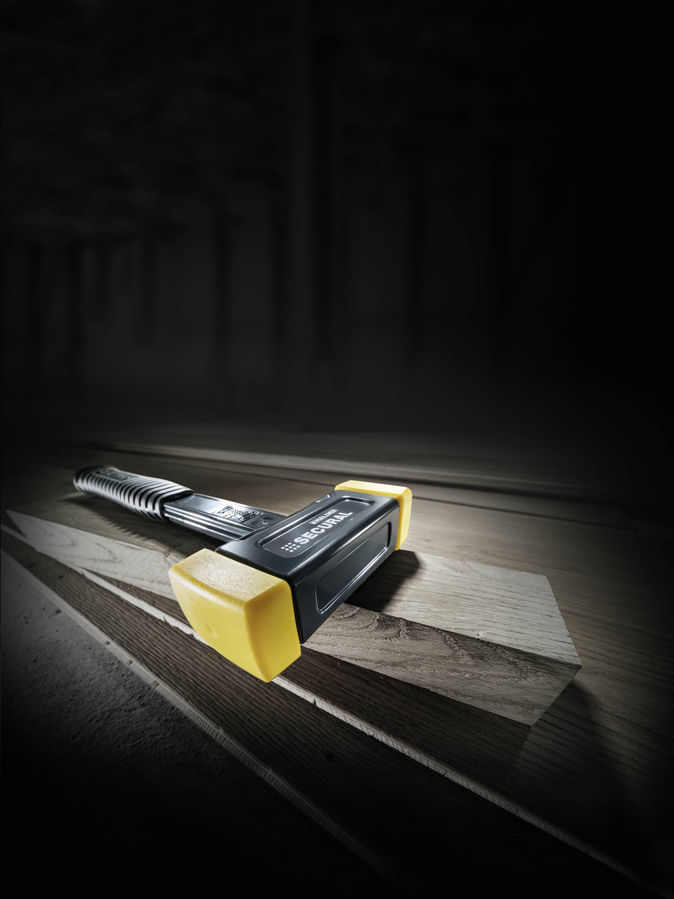 En hammer med gul gummibelægning ligger på træbrædder i et svagt oplyst rum.