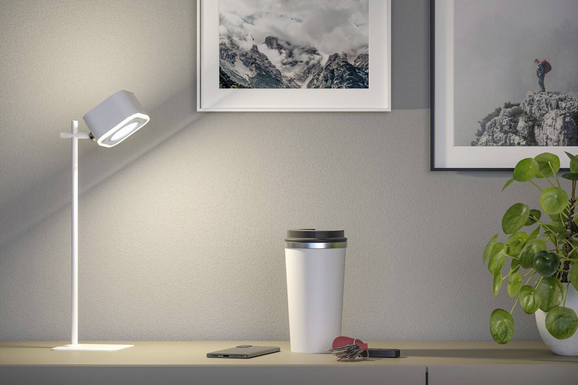 Et skrivebord med en lampe, kaffekop, smartphone og nøgler ovenpå. På væggen hænger billeder, en plante til højre.