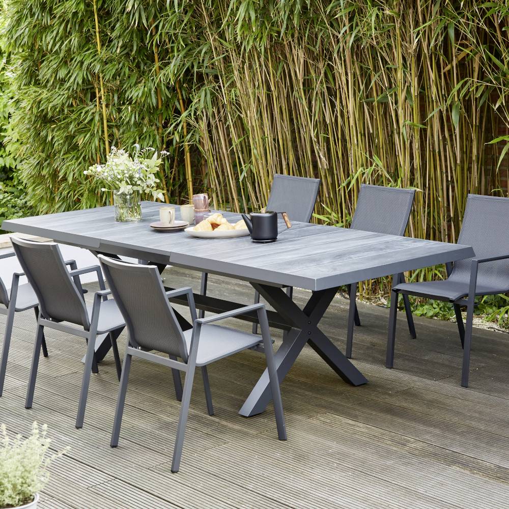 Et moderne havebord i træ, dækket med service og en gryde, på en terrasse med fem stole og bambus-planter i baggrunden.