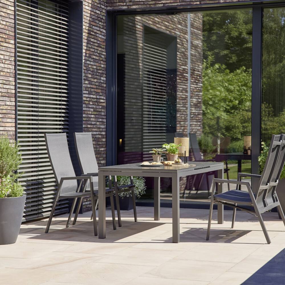 En moderne have med et bord, fire stole og planter i potter på en terrasse. Store glasvinduer i baggrunden.