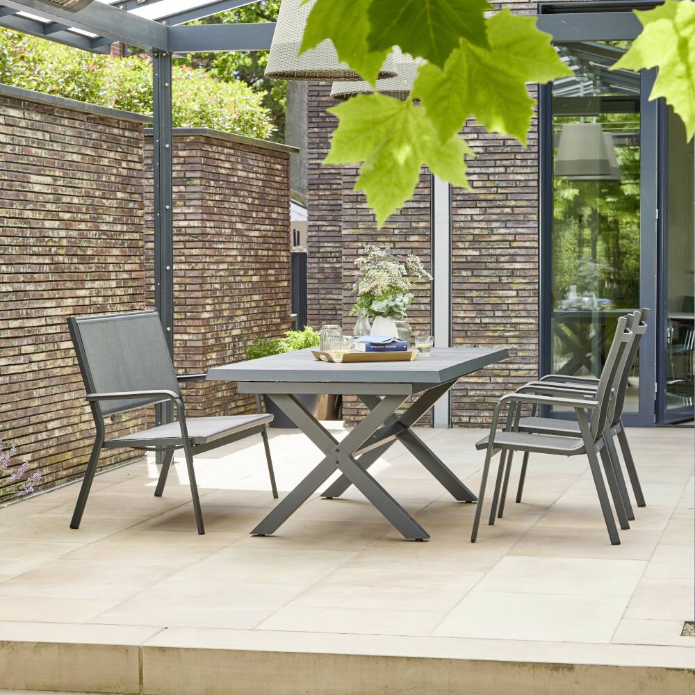 Terrasse med to grå metalstole og et bord. I baggrunden en murstensmur og glasvindue, omgivet af planter.