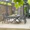Terrasse med moderne grå metalstole og -bord i en have med murede vægge og glastag, omgivet af grøn vegetation.
