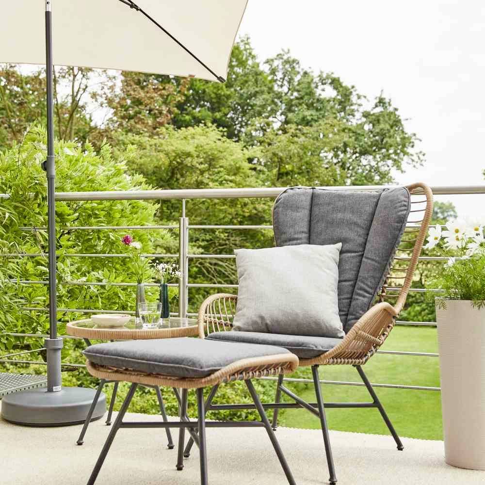 En rattanløungestol med puder på en altan, omgivet af grønne planter og en parasol, skaber en afslappet udendørs atmosfære.