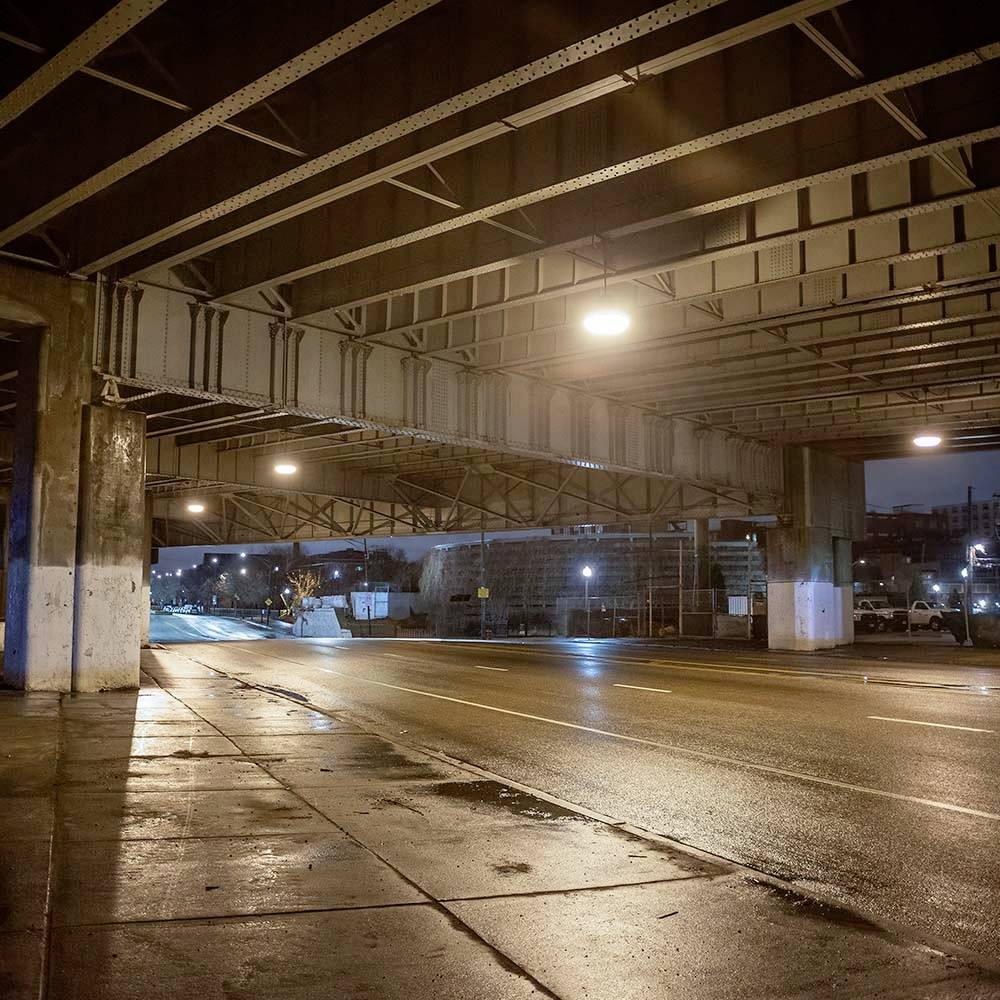 En tom, fugtig vejtunnel om natten, oplyst af gadelamper; urban omgivelse synlig i baggrunden.