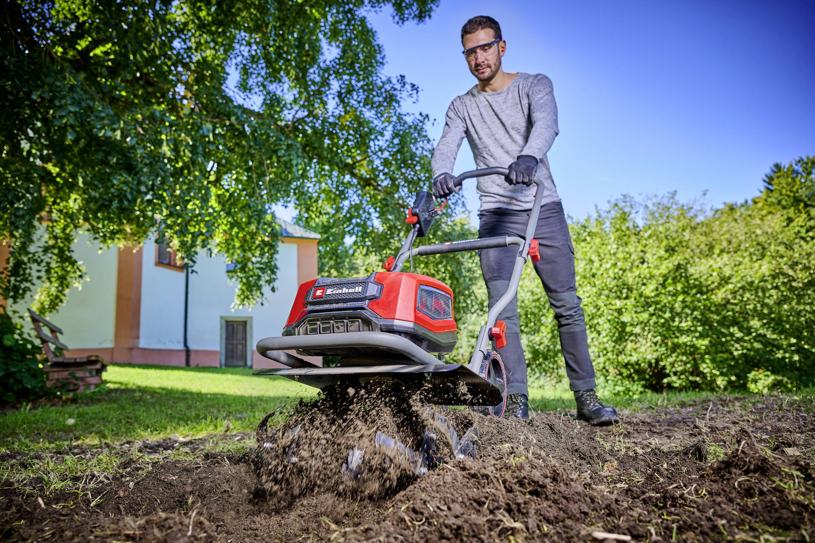 En mand betjener en elektrisk havekultivator på en grøn græsplæne foran et hus, idet han bærer beskyttelsesbriller og handsker.