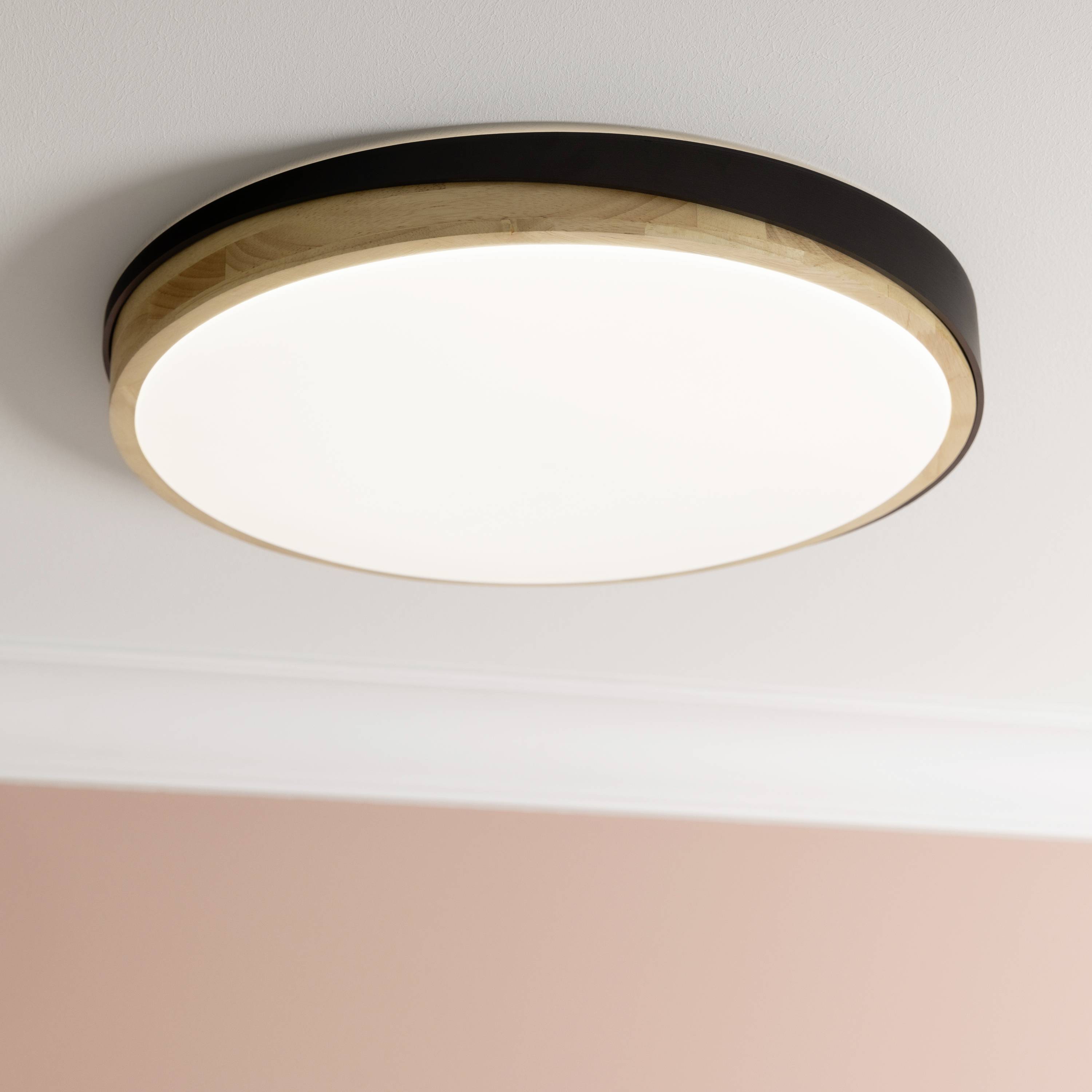 En rund loftslampe i lyst træ, monteret på en hvid lofts. Den lyser jævnt og giver rummet en behagelig lysfylde.