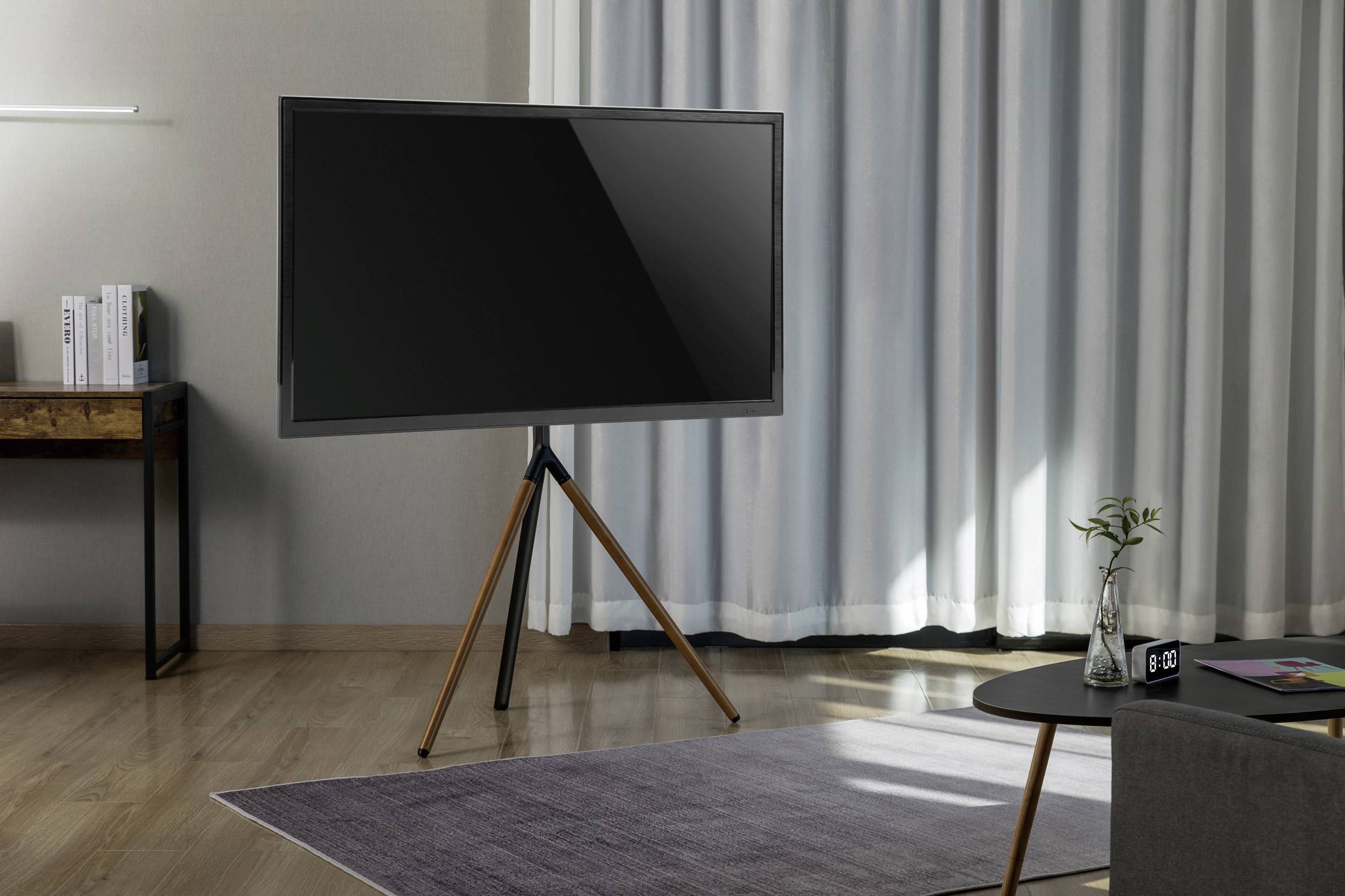 Stue med moderne fladskærms-tv på stativ, trægulv, grå gulvtæppe, skænk med bøger, bord med vase og gardiner i baggrunden.