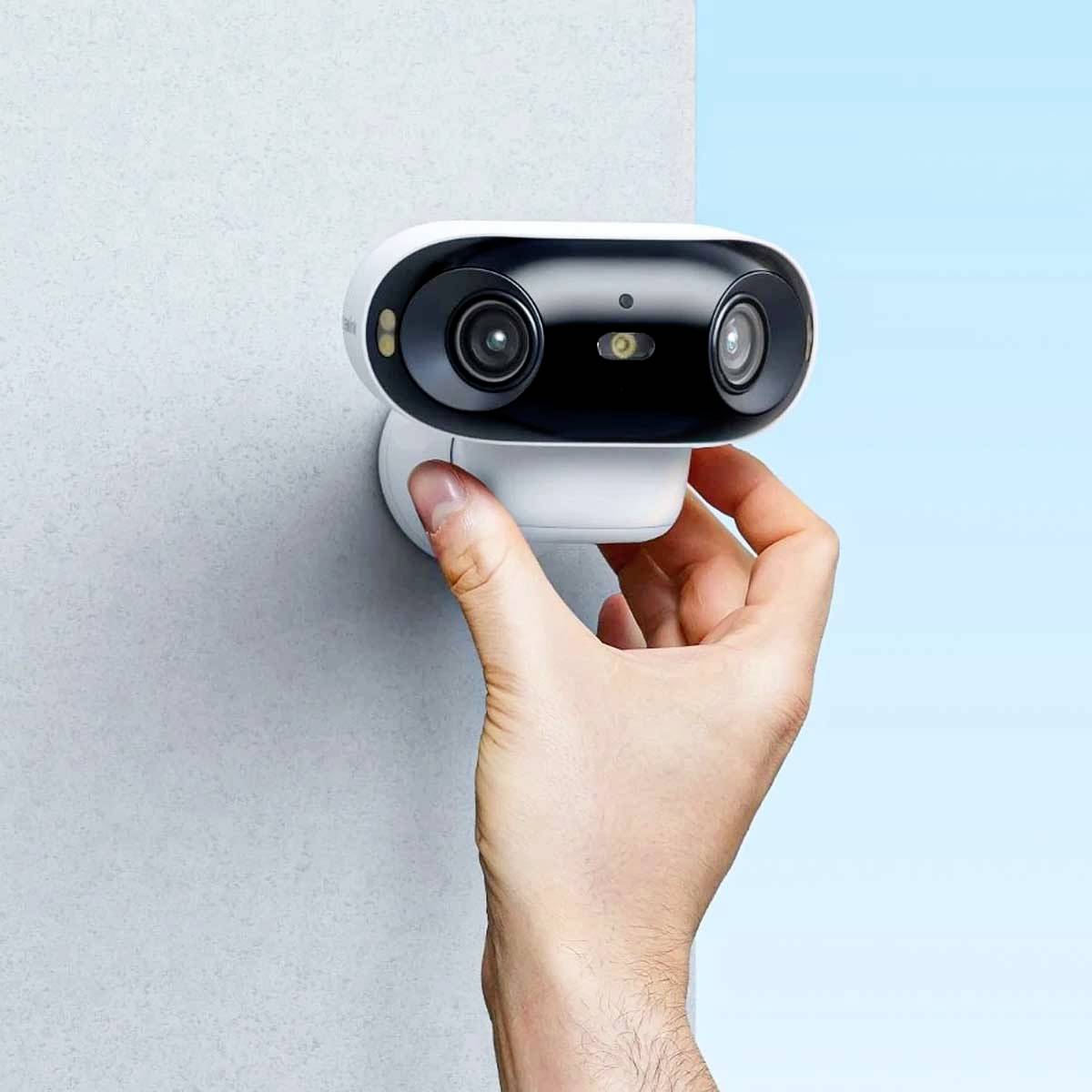 En hånd holder et lille, todelt sikkerhedskamera, der er monteret på en væg, foran en blå baggrund.