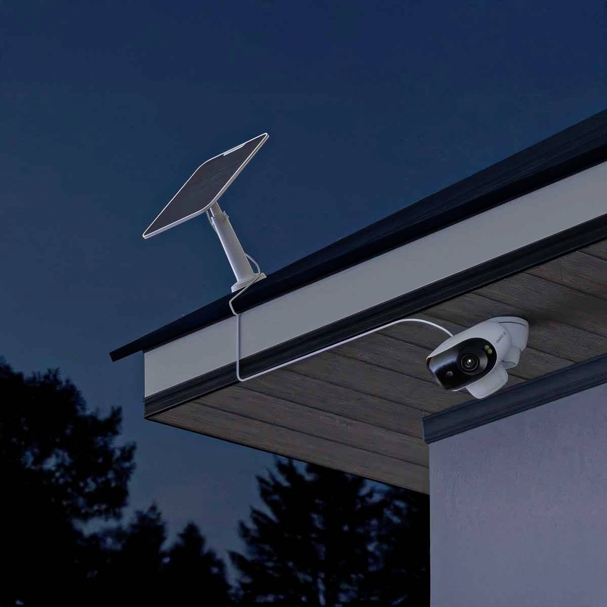 Solarpanel og overvågningskamera monteret på et hustag ved skumringstid, omgivet af træer.