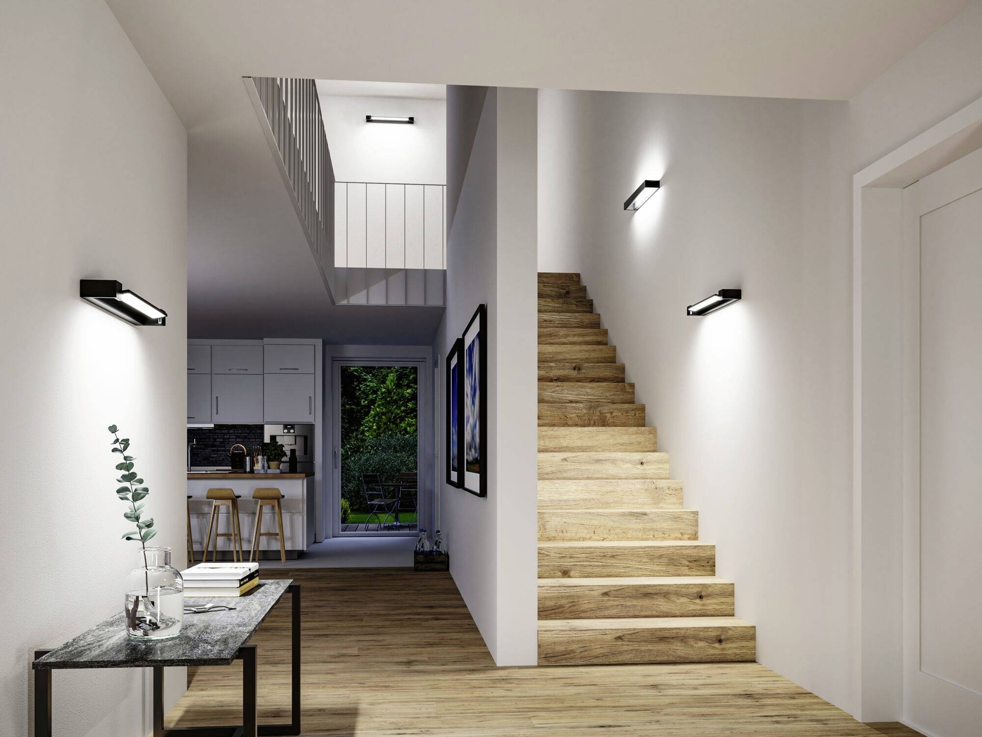 Entréparti med trægulv, trappe op til øverste etage, billeder på væggen og udsigt ind i et oplyst køkken i baggrunden.