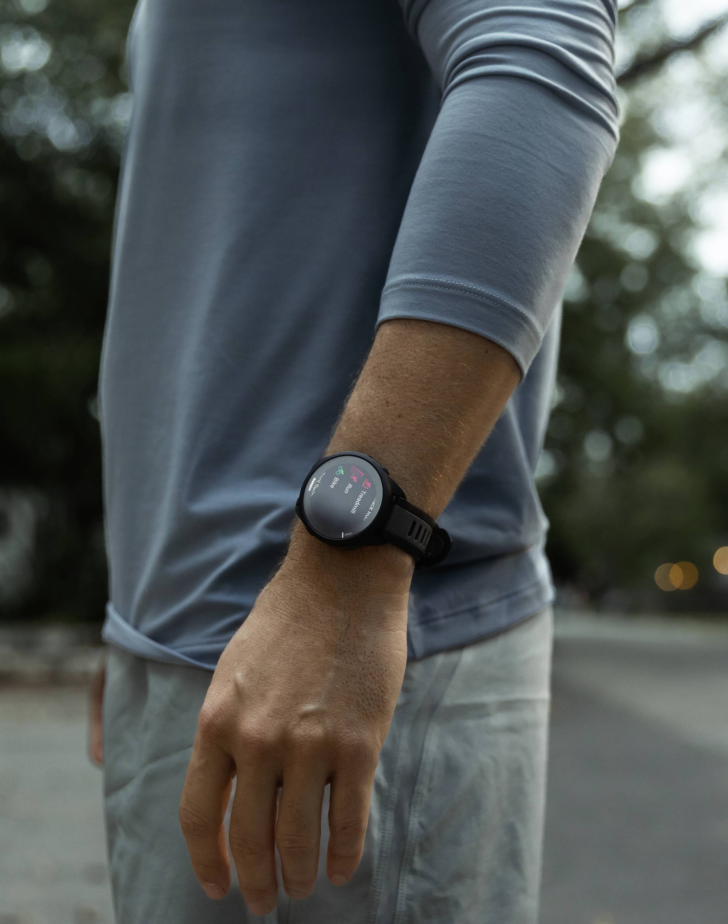 En person bærer en mørk smartwatch på deres venstre håndled. Personen står udendørs og har en lys langærmet trøje på.