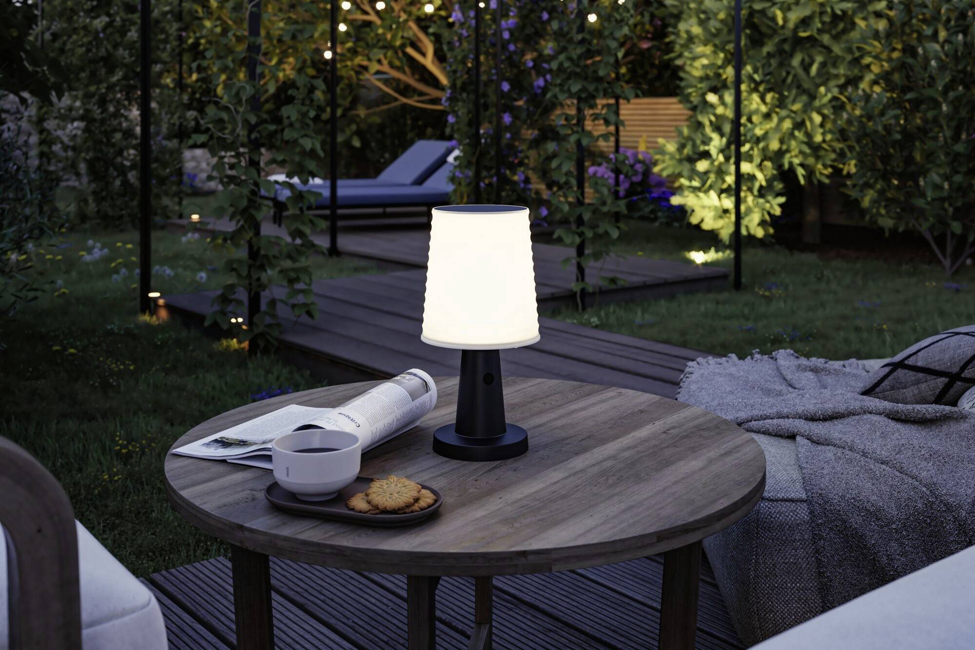 En hyggelig udendørs område om natten med et rundt bord, hvorpå der er en lampe, en kop og småkager. I baggrunden en oplyst have.