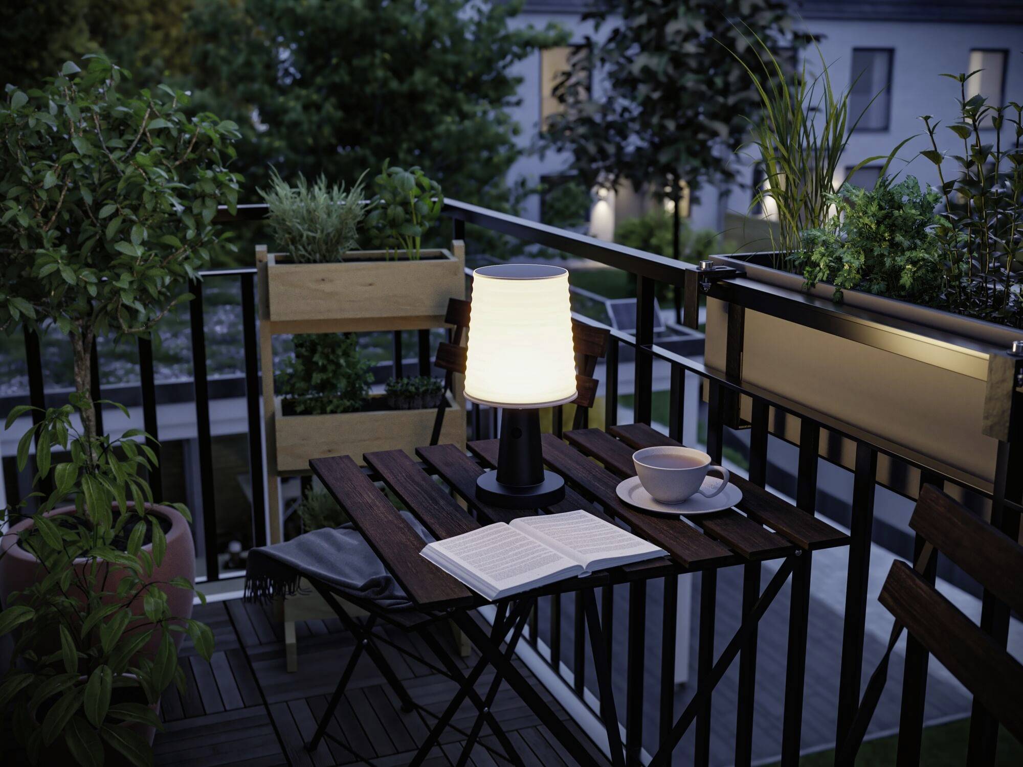 Balkonen viser et bord med en lampe, en åben bog og en kop, omgivet af planter og aftenlys.