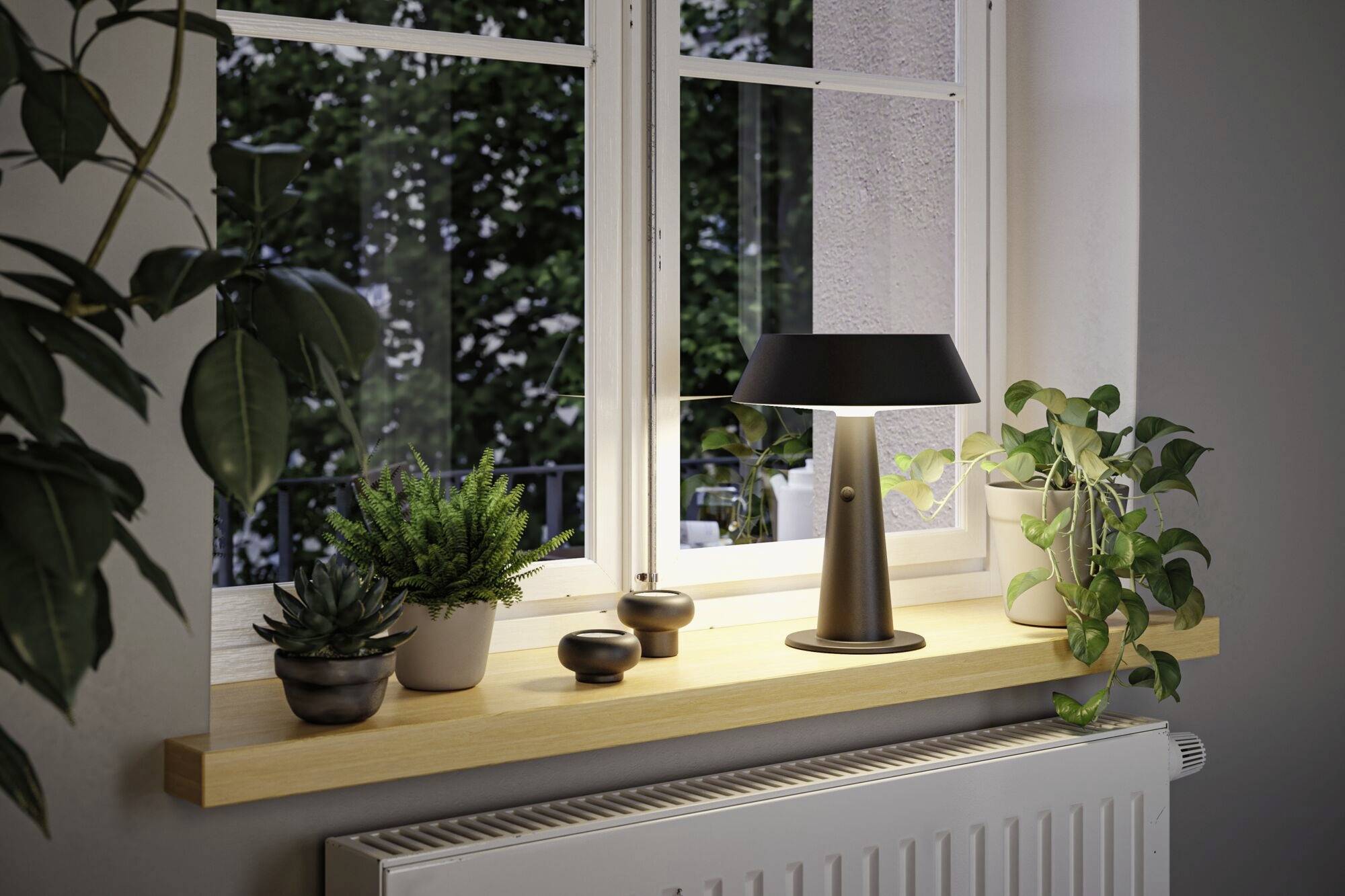 Et vinduesparti med flere planter i potter og en moderne bordlampe, bag hvilket der er udsigt til grønne træer udenfor.