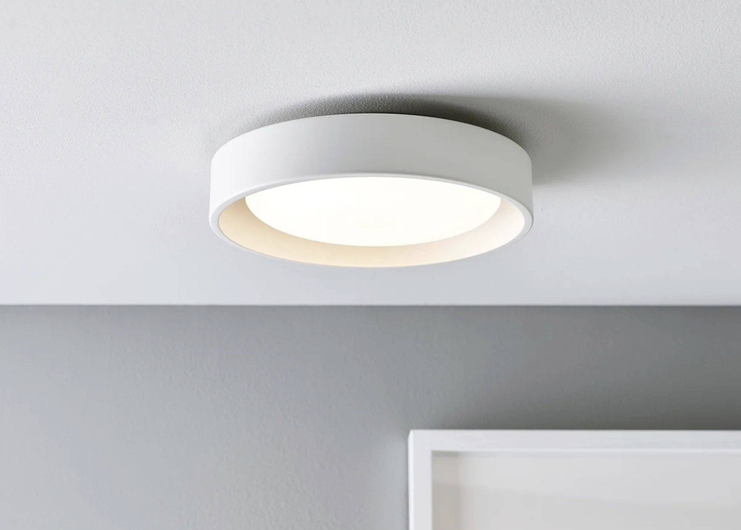 En rund, moderne loftslampe i hvidt metal udsender blidt, diffust lys i et minimalistisk indrettet rum.