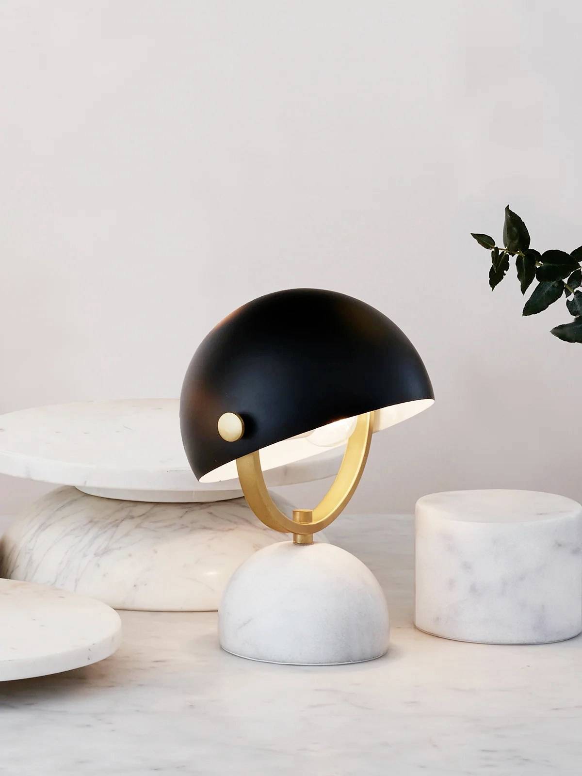 Moderne bordlampe med buet, sort skærm og guldfarvet sokkel placeret på hvidt marmorბord, dekoreret med planter.