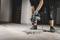 En person borer med en elektrisk slagboremaskine i et betongulv. Støvpartikler spredes omkring borepunktet.