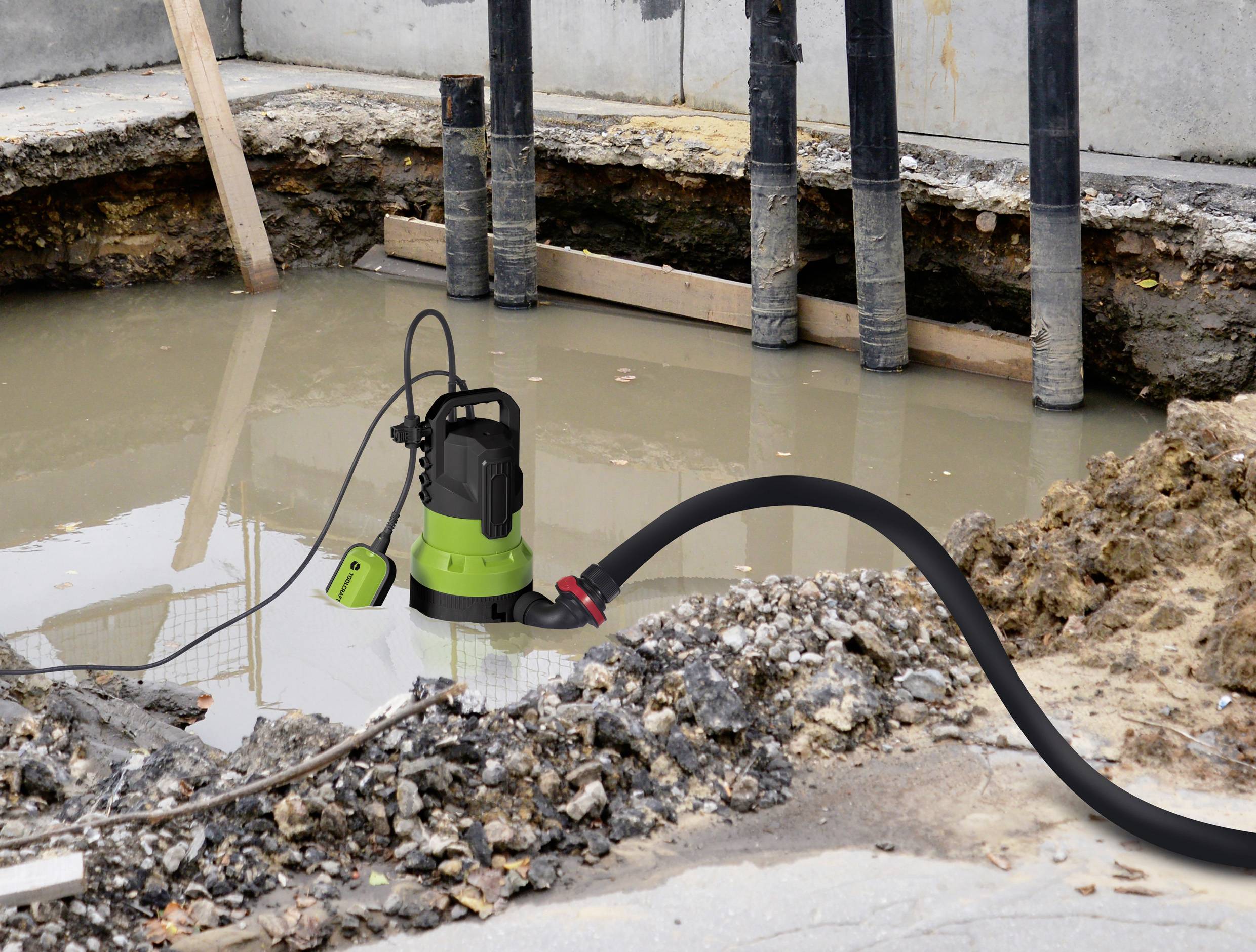 En grøn dykpumpe afvander en byggeplads med mudret vand. Sort slange fører væk fra pumpen.