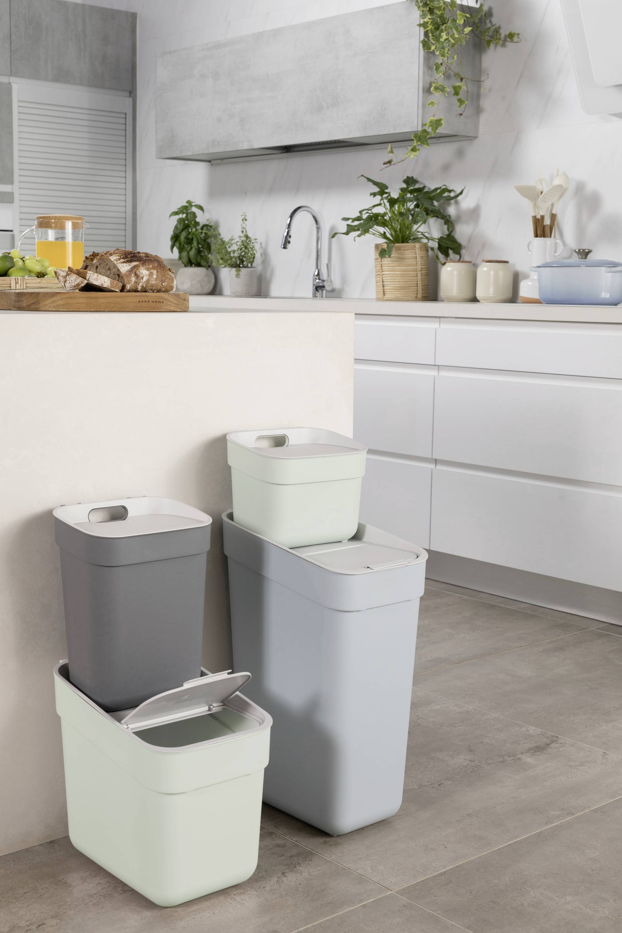 Tre skraldespande i forskellige farver står i et moderne køkken ved siden af en bordplade med en karaffel og bagværk.
