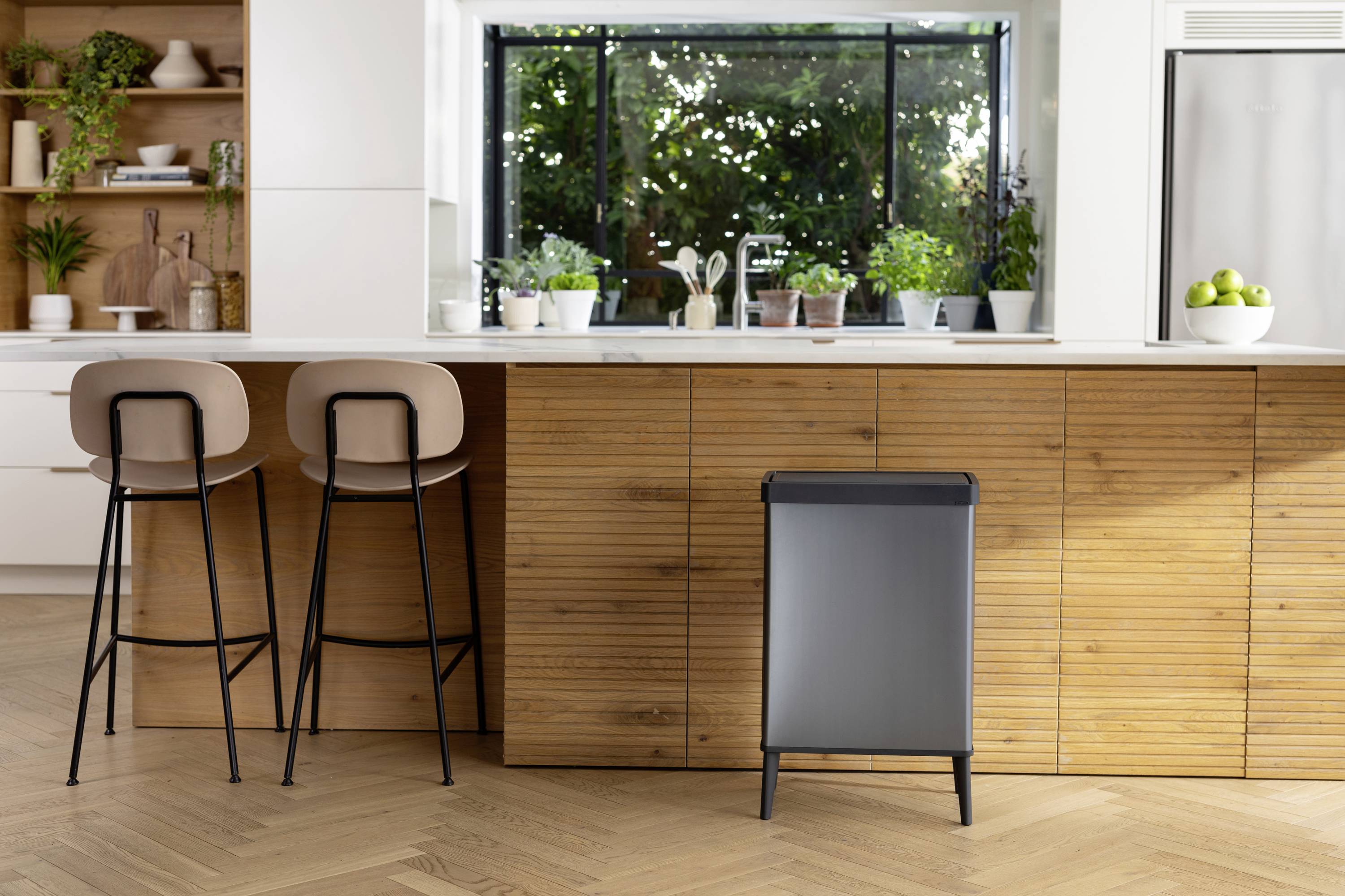 Moderne køkken med træelementer og planter på bordpladen. To barstole står ved en bardisk, med en skraldespand foran.