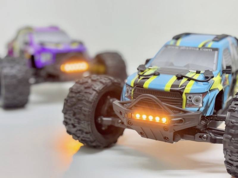 To fernbetjente monstertrucks, én blå-gul og den anden lilla-sort, kører på en hvid overflade med lysende forlygter.