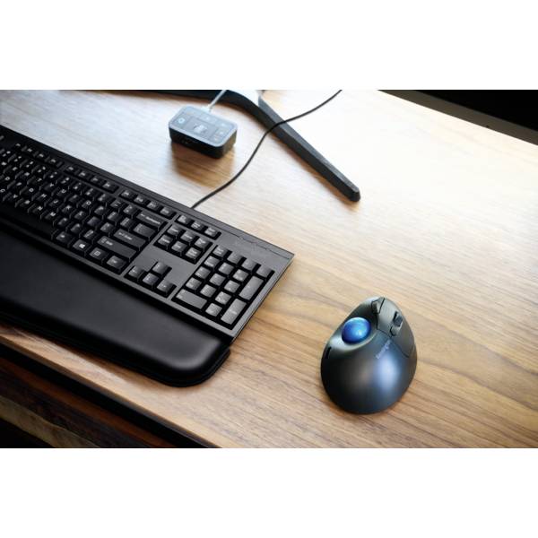 Et computertastatur med håndledsstøtte på et træbord, ved siden af en ergonomisk mus med en blå trackball-kugle.