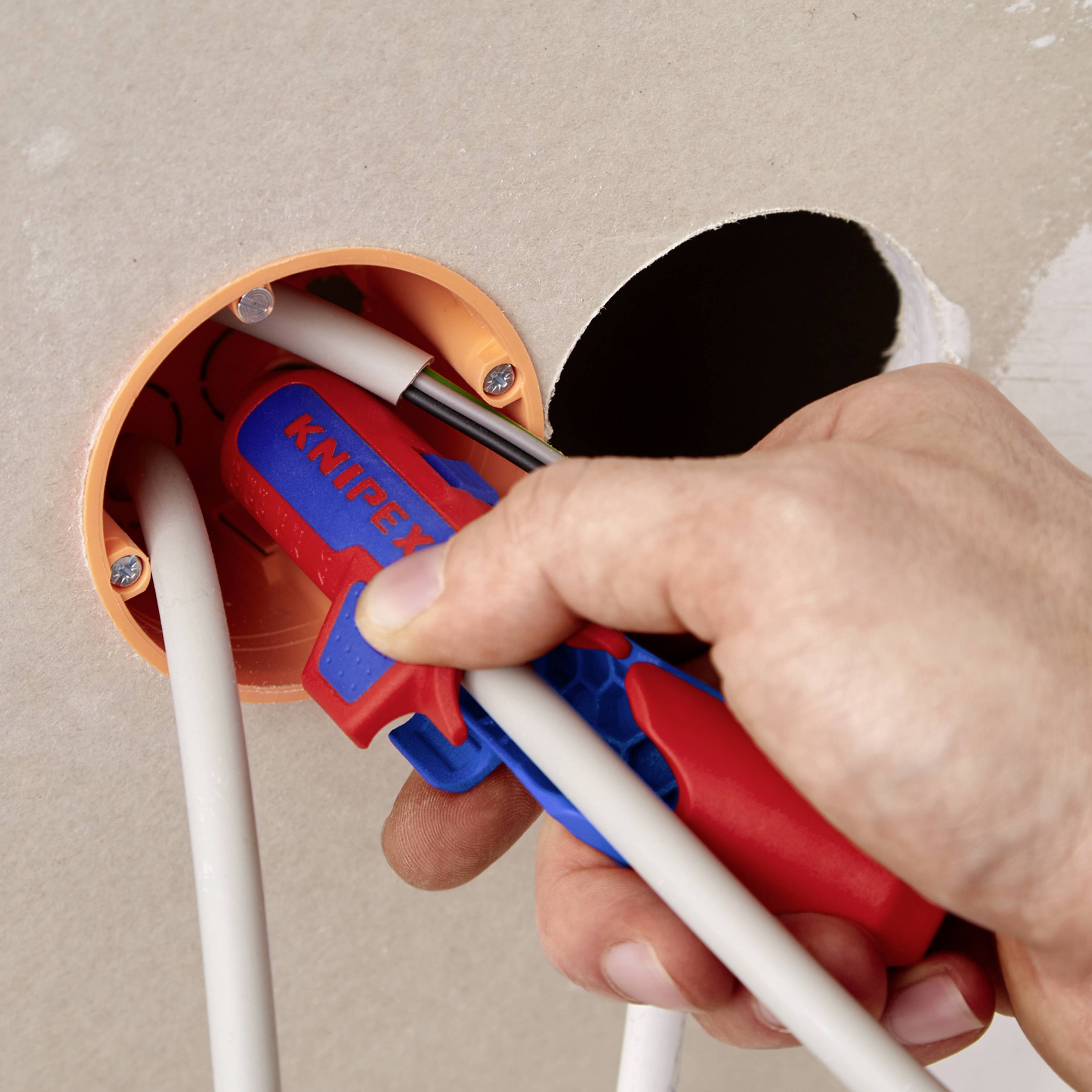 En hånd holder et Knipex-værktøj og klipper en ledning inde i en vægåbning.