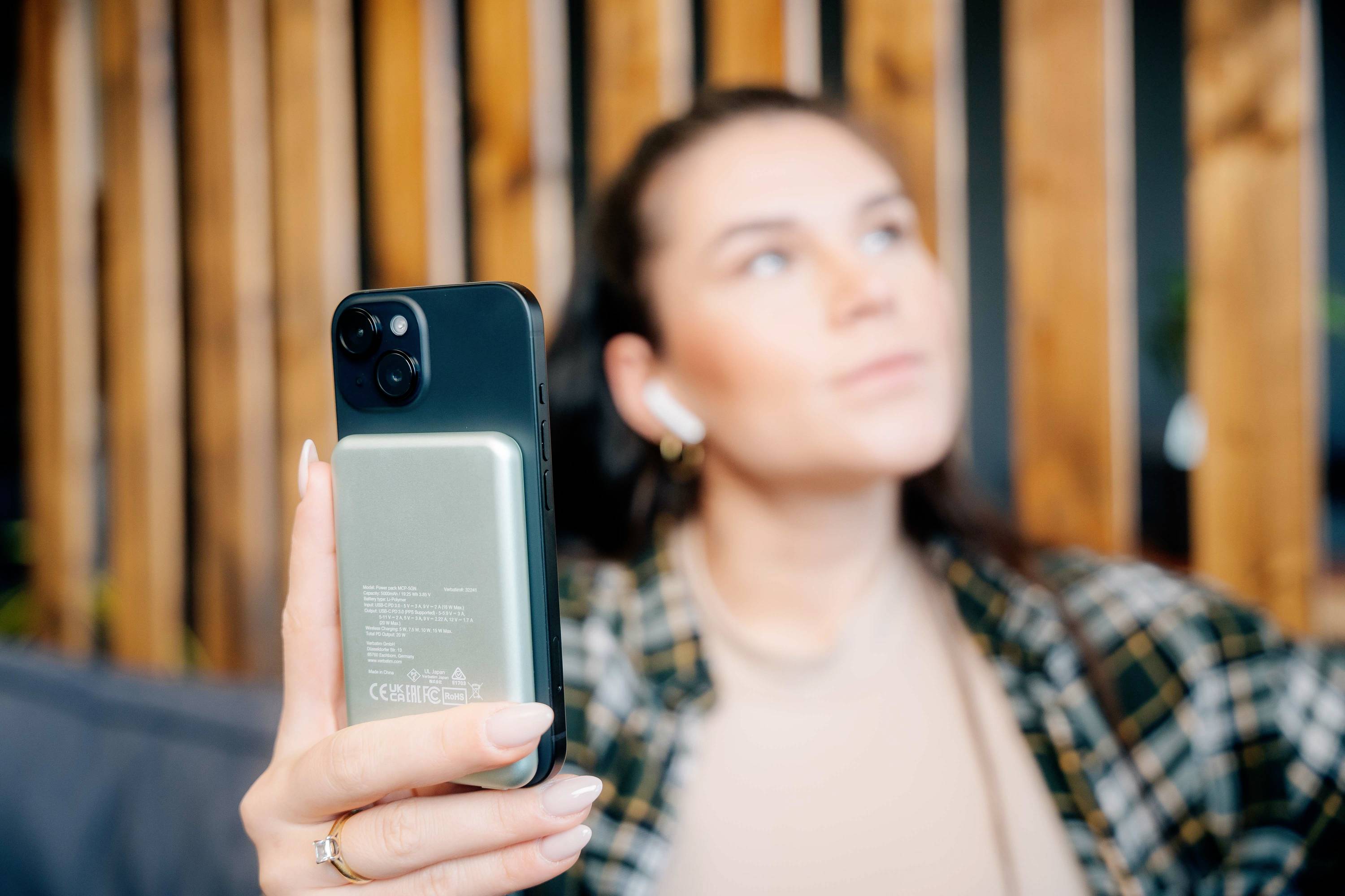 En person holder en smartphone, kigger op og bærer trådløse høretelefoner. I baggrunden er en træpanel.