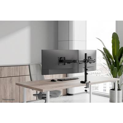 To monitorer står på et skrivebord på en justerbar monitorarm foran et vindue, ved siden af en plante, i et moderne kontormiljø.