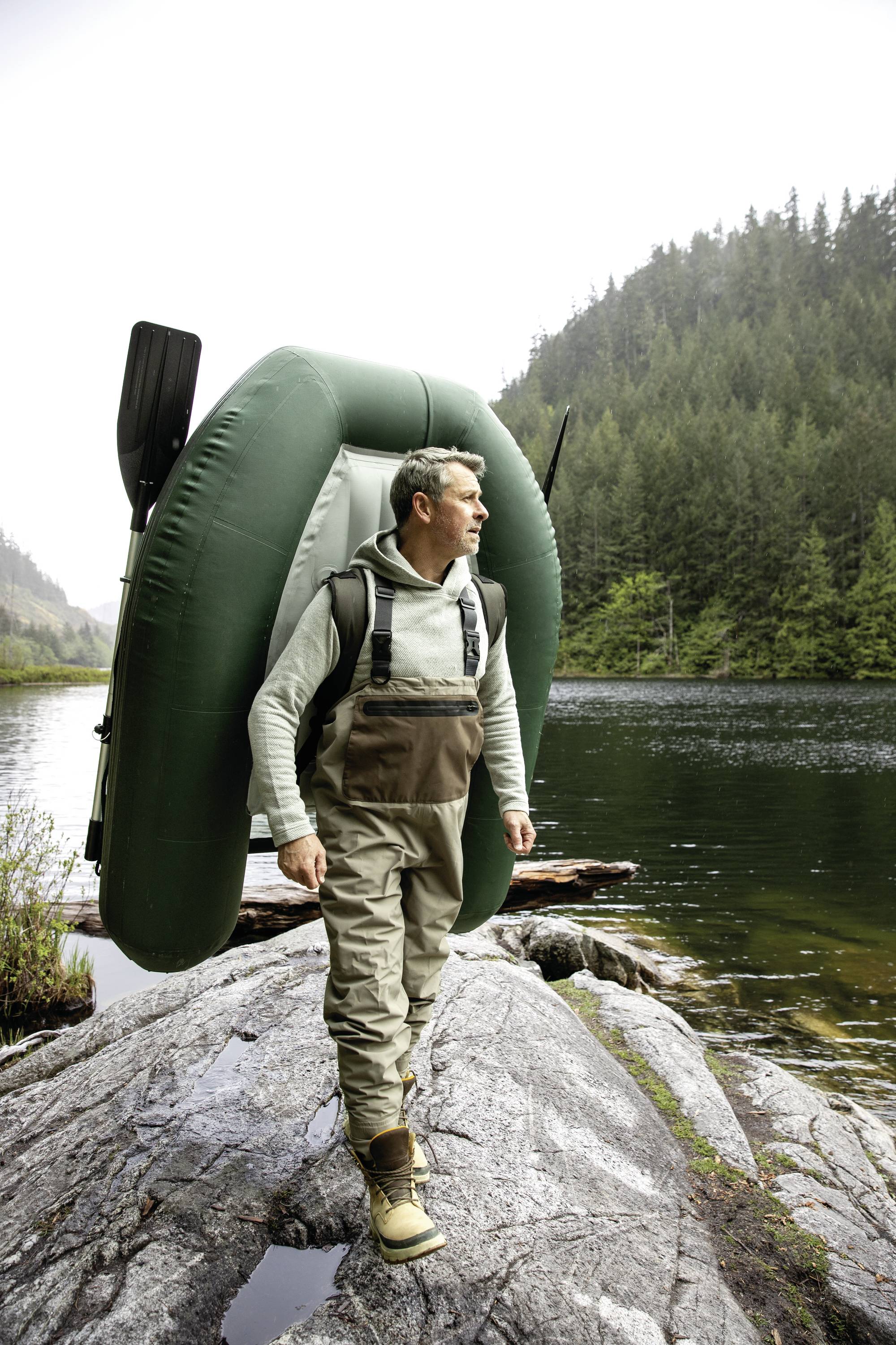 En mand i outdoor-tøj bærer en oppustelig båd på ryggen og står på klippefuld grund ved en skovklædt søbred.