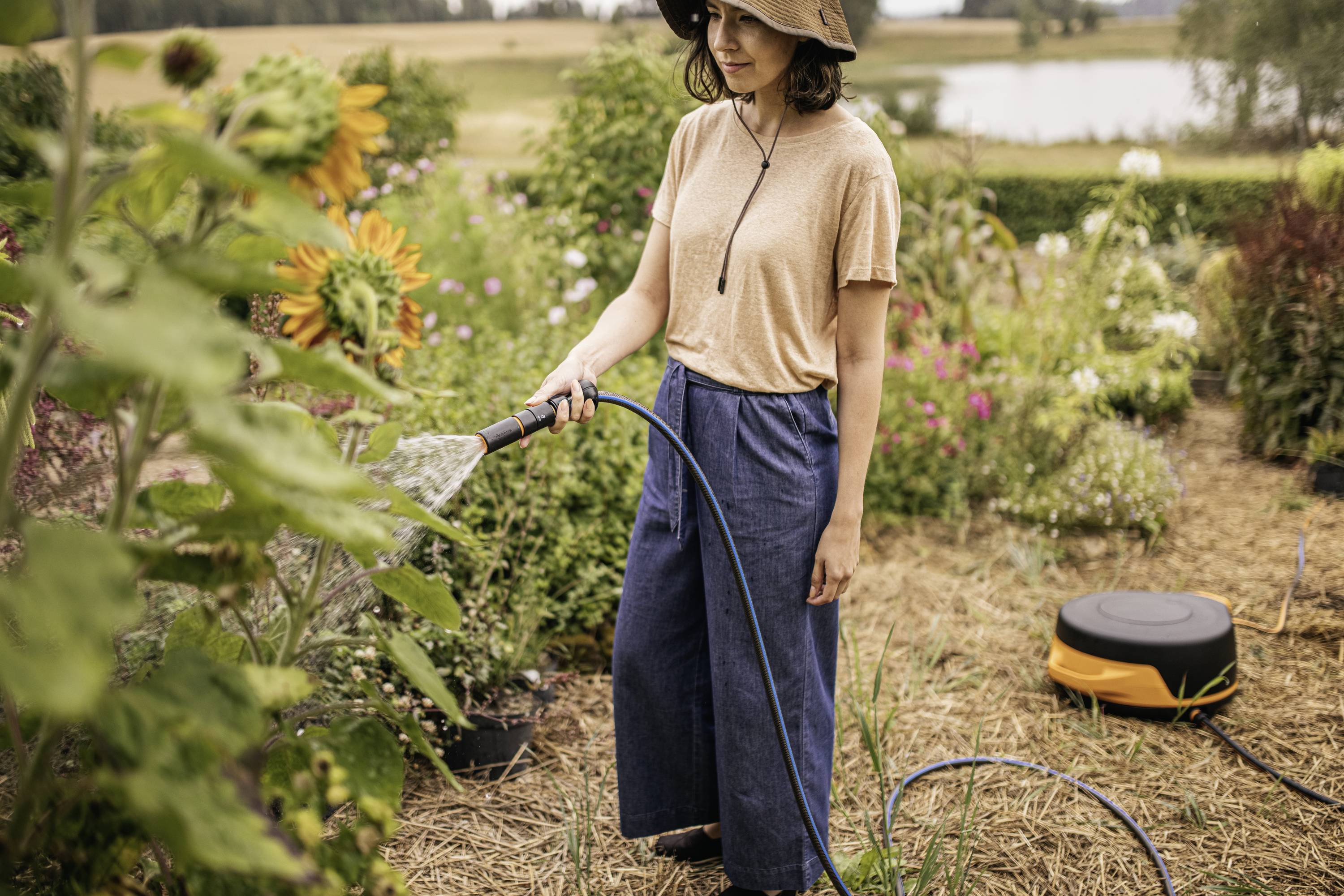 En person med en hat vander planter med en haveslange i en blomstrende have. I baggrunden ses solsikker og yderligere blomsterbede.