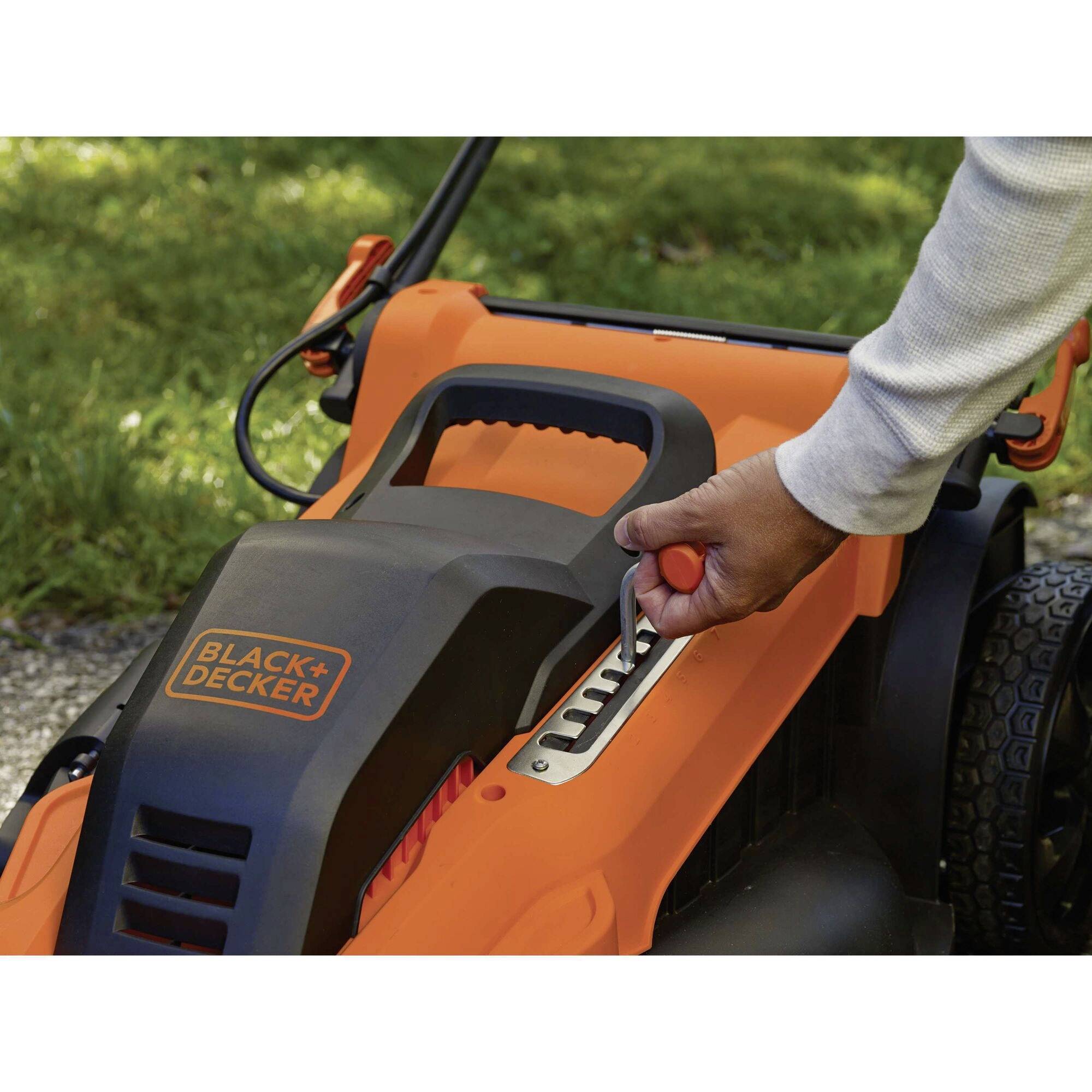 En hånd justerer klippehøjden på en orange plæneklipper med påskriften 'Black+Decker'.