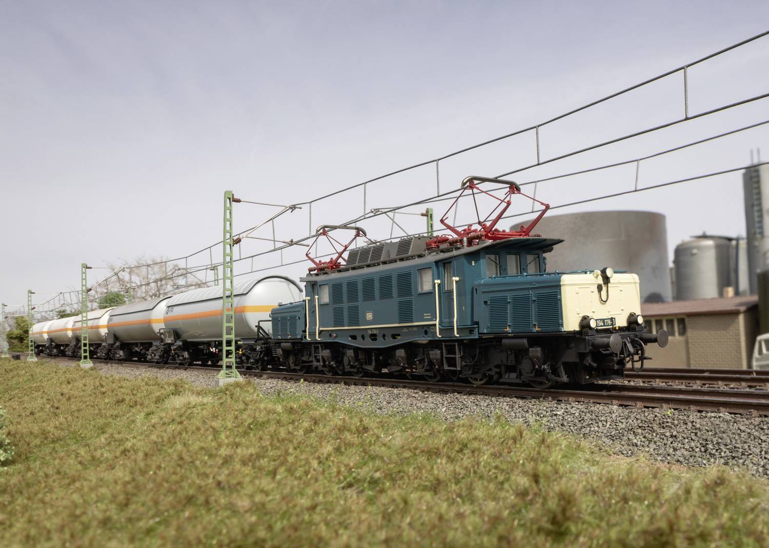 En modeltog med en blå og beige lokomotivfører trækker sølvfarvede tankvogne på en miniature-jernbanestrækning udendørs.